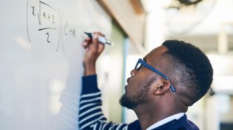 Young man writing on a whiteboard in a classroom Man with glasses in striped jumper writing mathematical equation on whiteboard